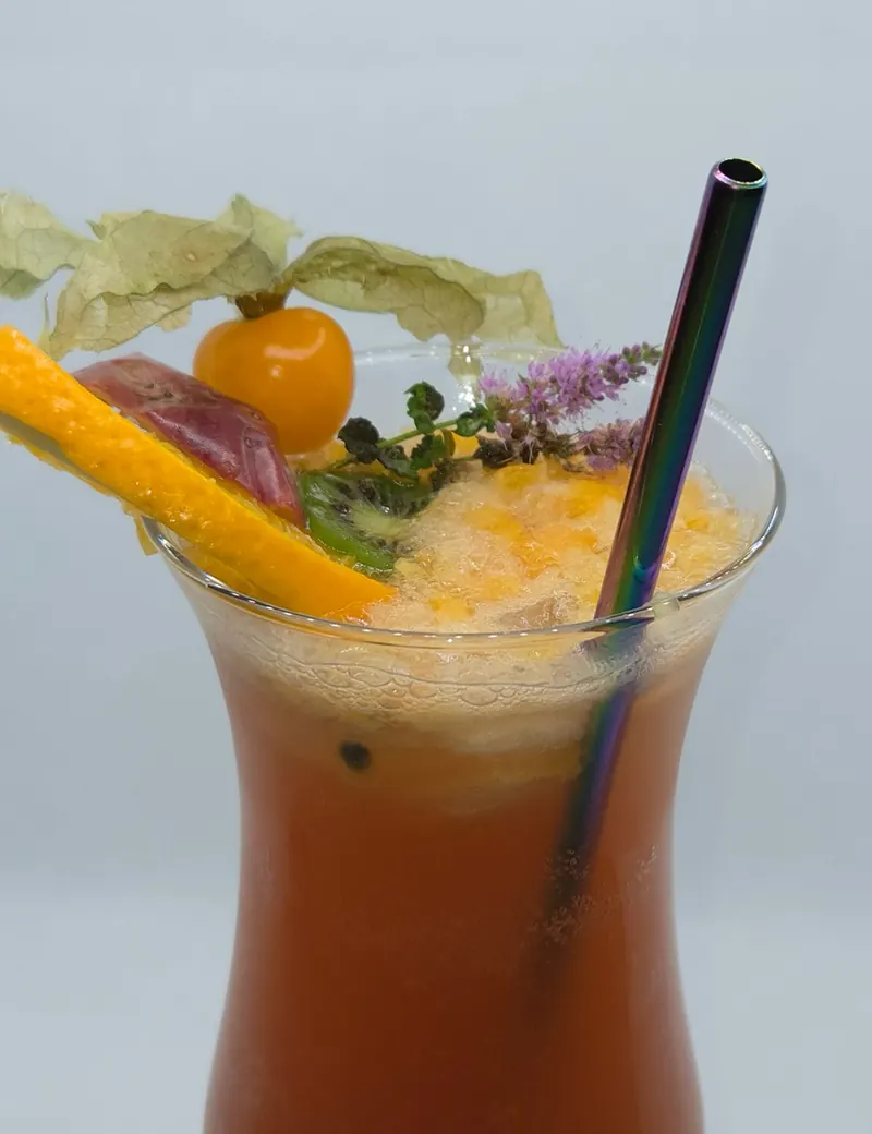 Orangefarbener Cocktail mit Physalis, Orangenzeste und Kräutern, dazu ein Trinkhalm aus Metall