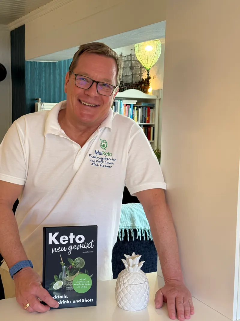 Maik Roemer lacht in die Kamera und hält sein Buch „Keto neu gemixt“ in der Hand