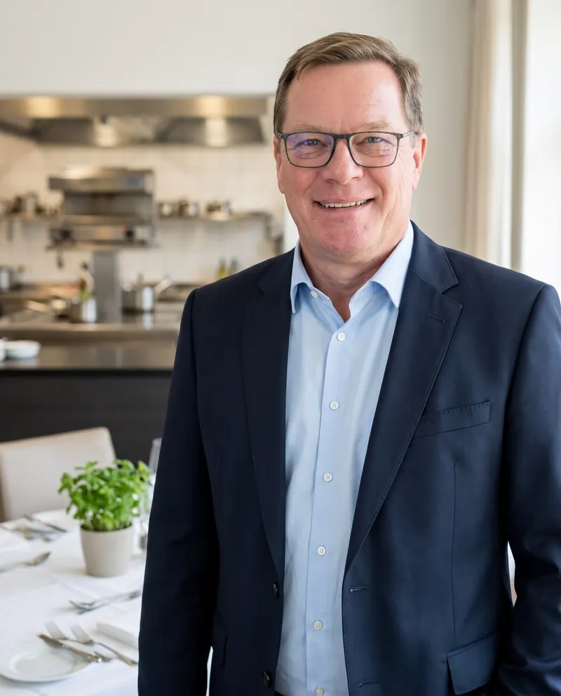 KI-generiertes Bild: Maik Roemer im dunkelblauen Anzug lächelt in einem hellen Restaurant, im Hintergrund eine offene Profiküche