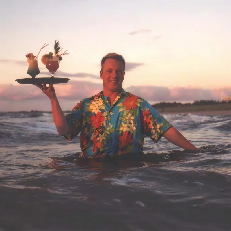 Altes Foto, mit KI bearbeitet: Maik Roemer steht im Hawaiihemd hüfttief im Meer und hält im Abendlicht ein Tablett mit zwei Cocktails