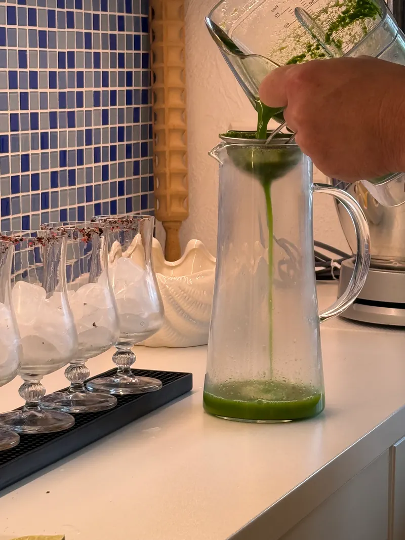 Grüner Kräutersaft läuft durch ein Sieb in eine Glaskaraffe